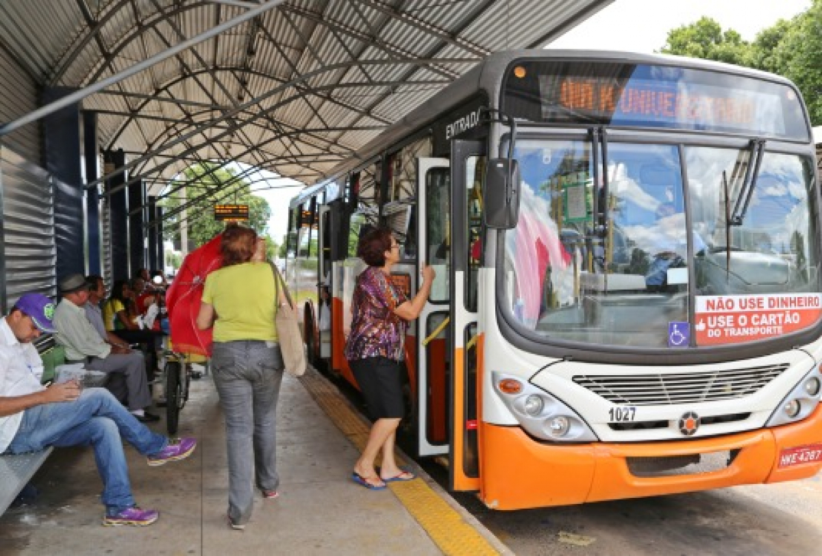 Passageiros embarcam em um ônibus urbano em terminal coberto, com aviso para uso exclusivo de cartão no transporte.