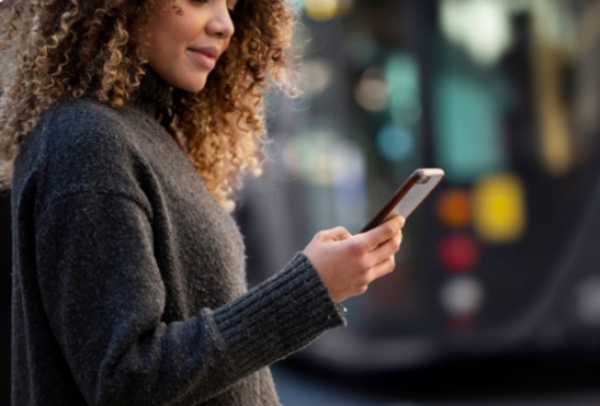 Mulher sorri enquanto usa o celular em frente a um ônibus urbano. 