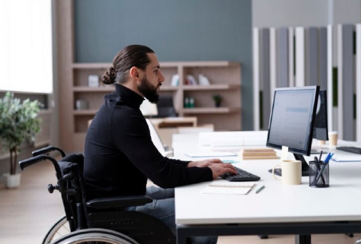 A imagem mostra uma pessoa sentada em uma cadeira de rodas, trabalhando em um computador em uma mesa de escritório.