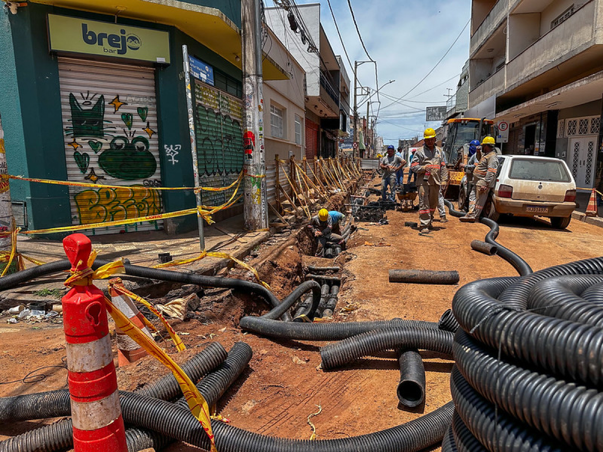 Obras de infraestrutura em via urbana, com trabalhadores instalando tubulações subterrâneas e área isolada para garantir a segurança.