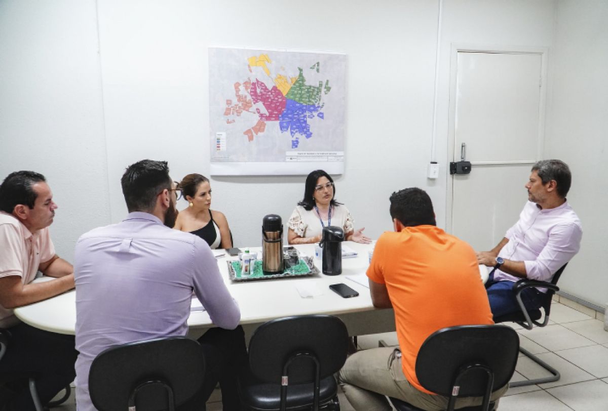 A imagem mostra um grupo de cinco pessoas reunidas em uma mesa de escritório, discutindo algo, com um mapa colorido na parede ao fundo.