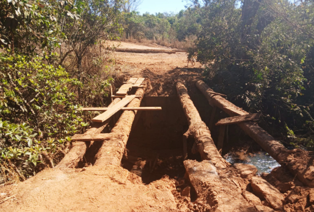 Ponte rústica de troncos e tábuas sobre um córrego em estrada de terra, em condições precárias.