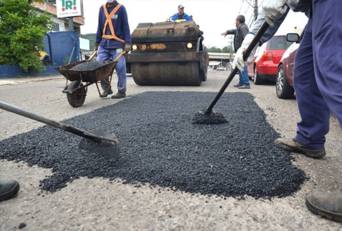 Trabalhadores realizam operação tapa-buracos aplicando e nivelando asfalto em via urbana.