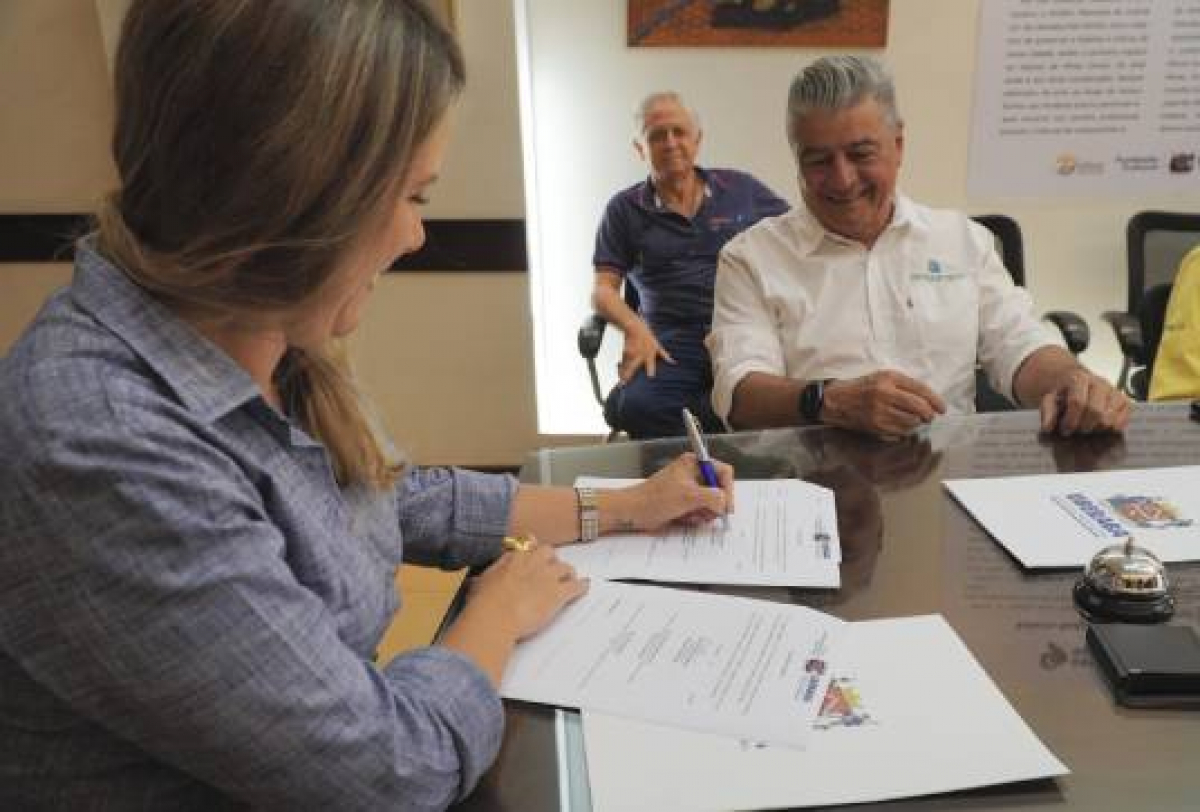 A imagem mostra uma pessoa assinando um documento em uma mesa, observada por várias outras pessoas ao fundo.