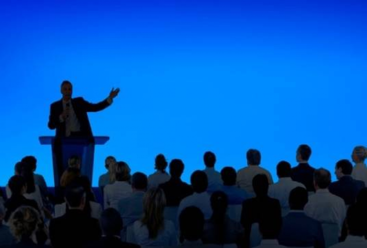A imagem mostra um palestrante em um palco falando para uma plateia, com um fundo azul vibrante.