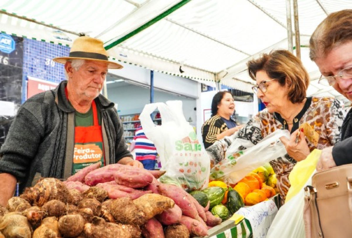 Feirante vende batatas e legumes frescos a clientes em uma feira livre.