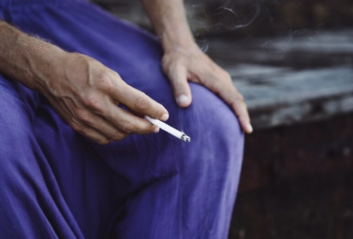 Pessoa sentada de roupa roxa segurando um cigarro aceso na mão.