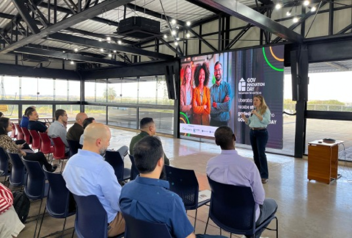 Palestrante apresenta sobre o evento GOV Innovation Day para um grupo de pessoas em auditório moderno.