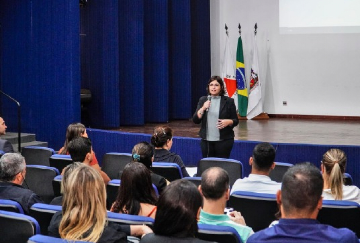 Mulher ministra palestra em auditório com plateia atenta e bandeiras ao fundo.