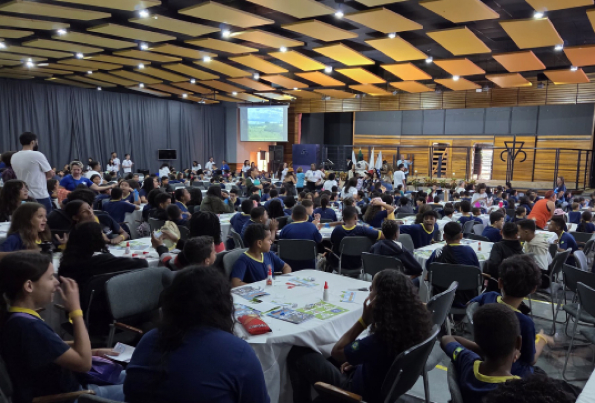 Evento educacional com grande participação de estudantes, realizado em um auditório, com mesas temáticas e apresentações projetadas.
