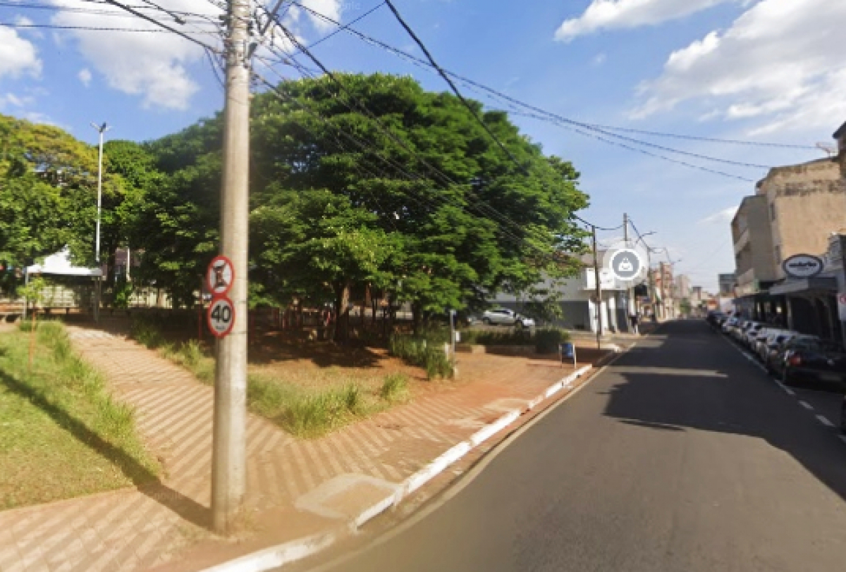 Rua urbana com praça arborizada, comércios ao lado e sinalização de trânsito visível.
