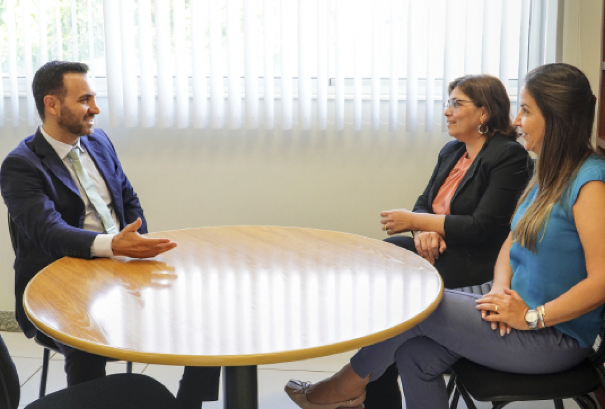 Três pessoas em uma reunião ao redor de uma mesa, conversando de forma profissional.