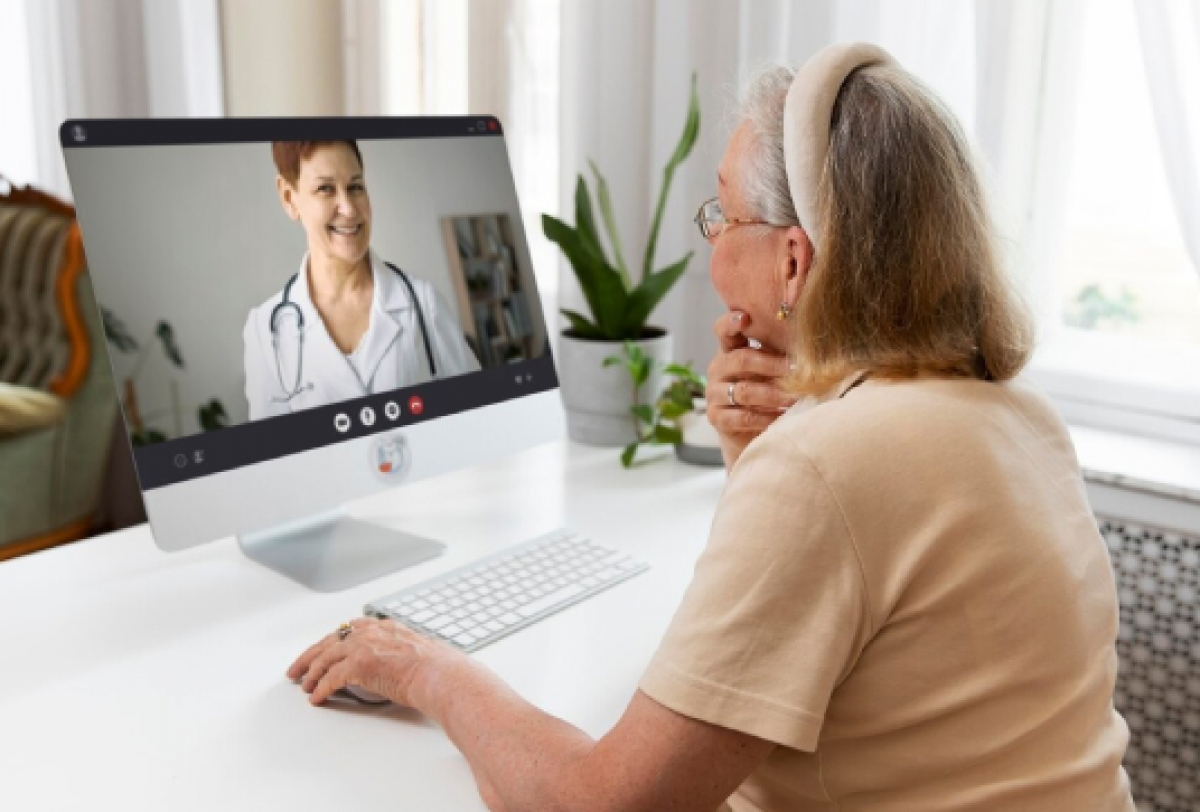 Idosa participa de uma consulta médica online, interagindo com uma profissional de saúde por videochamada em casa.