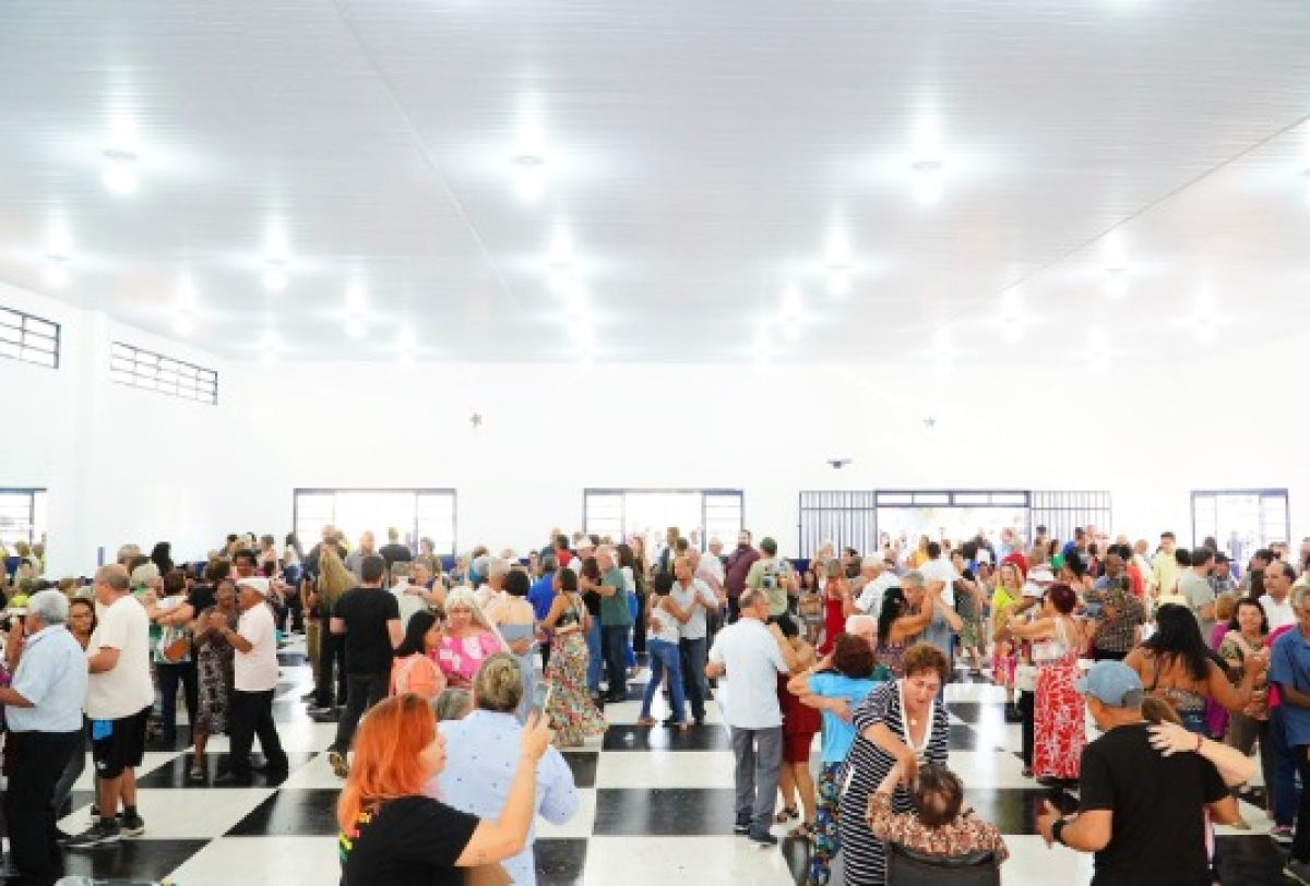 Grande grupo de pessoas participa de um baile ou confraternização em salão comunitário.