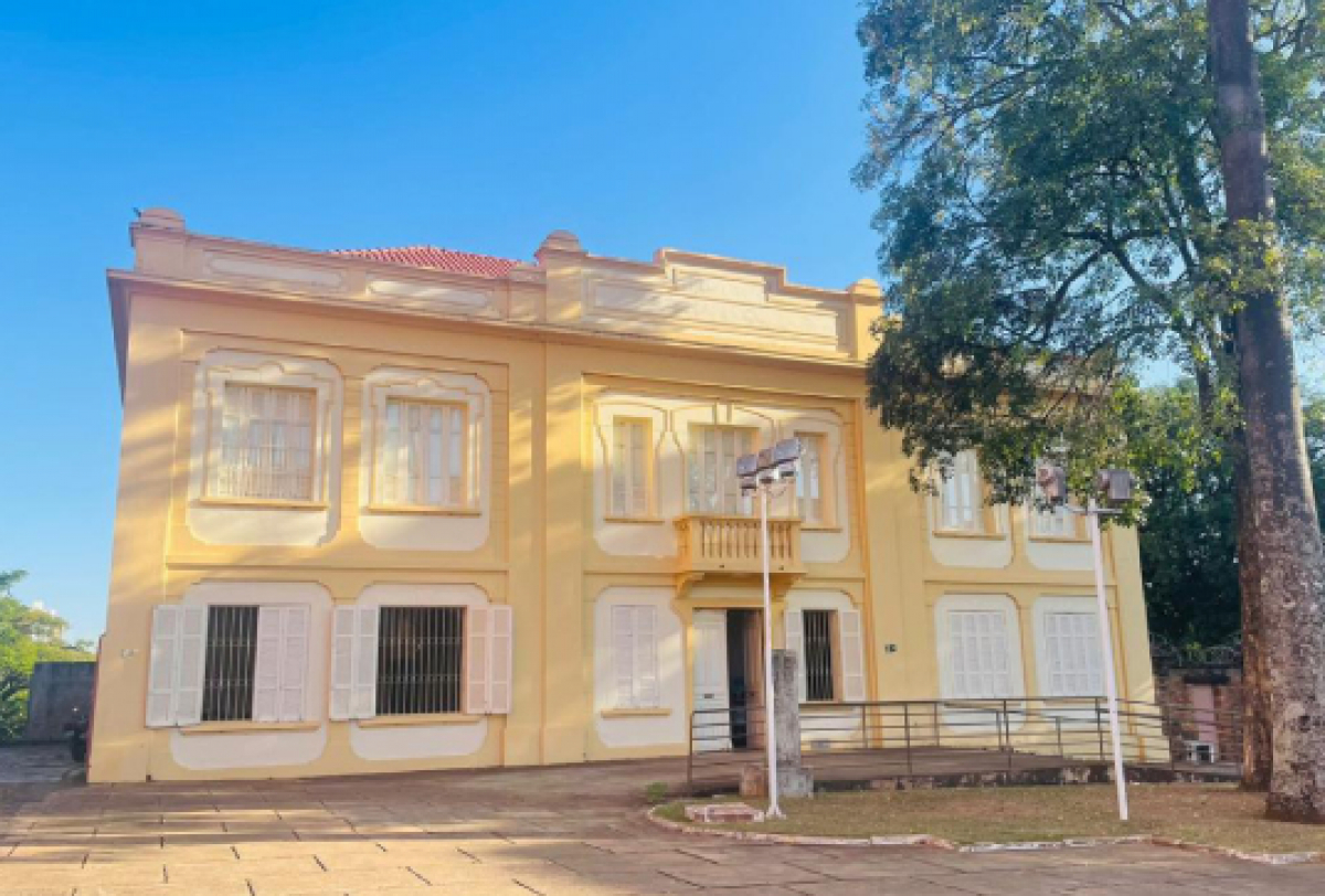 Prédio histórico de arquitetura clássica, em tons de amarelo, cercado por árvores e área pavimentada.