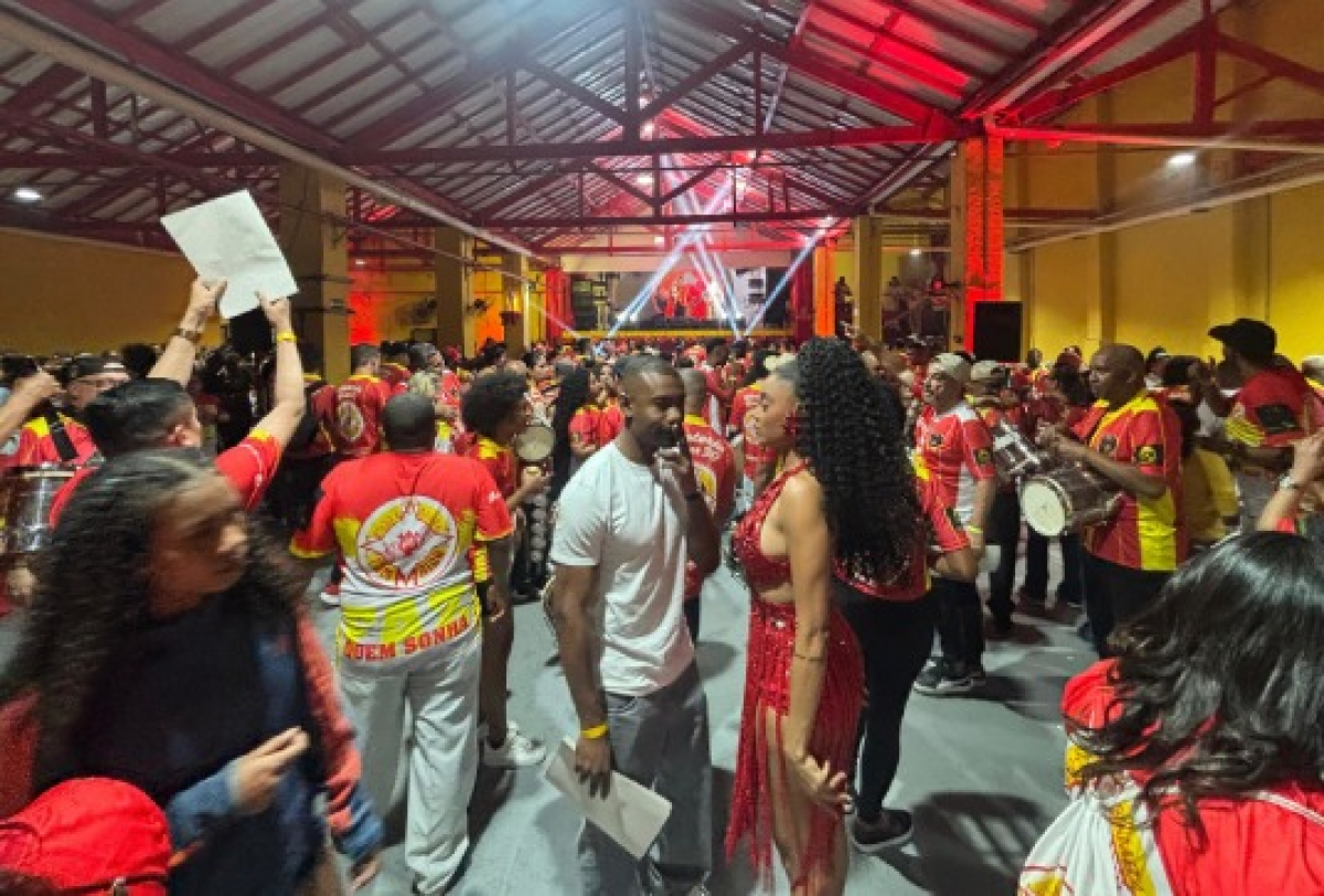 Ensaio animado de escola de samba reúne integrantes com fantasias e instrumentos em clima festivo.
