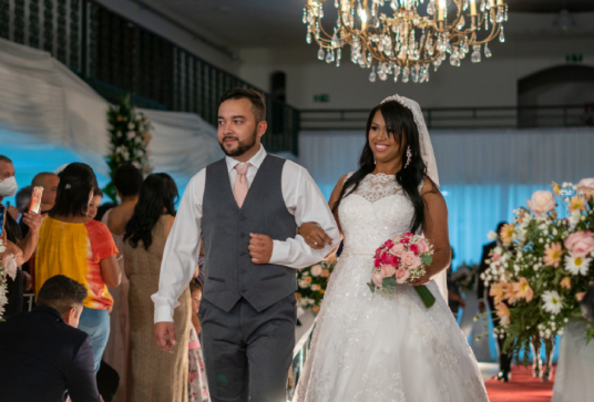 Uma noiva radiante e um noivo elegante caminham juntos em um corredor decorado com flores e um belo lustre.