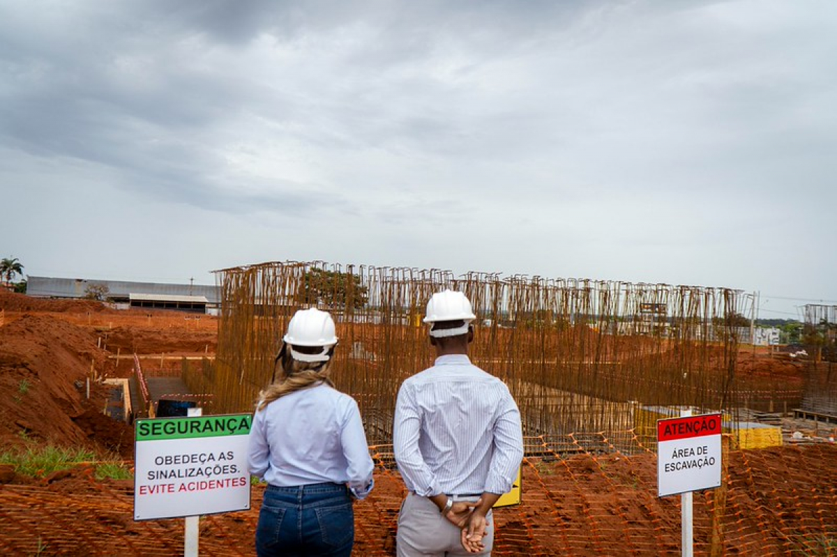 Duas pessoas usando capacetes observam uma obra de construção, cercada por placas de segurança e sinalização de área de escavação.
