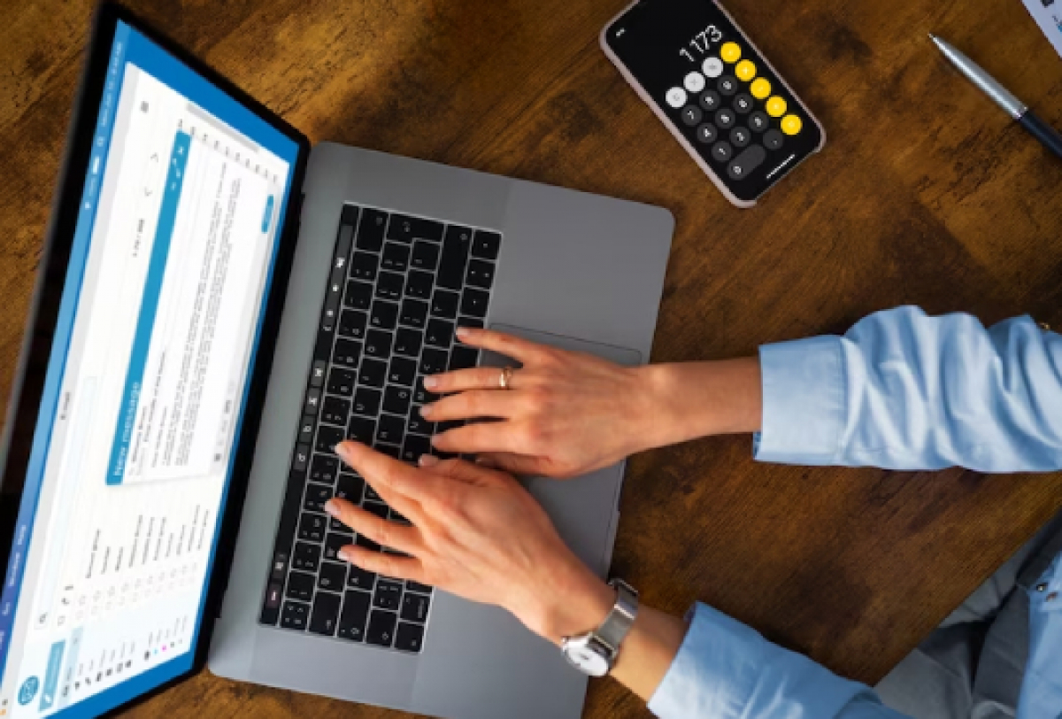 Pessoa digitando em um notebook sobre uma mesa de madeira, com calculadora e caneta ao lado.