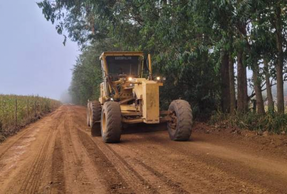 Máquina motoniveladora recupera estrada de terra em área rural cercada por árvores e plantações.