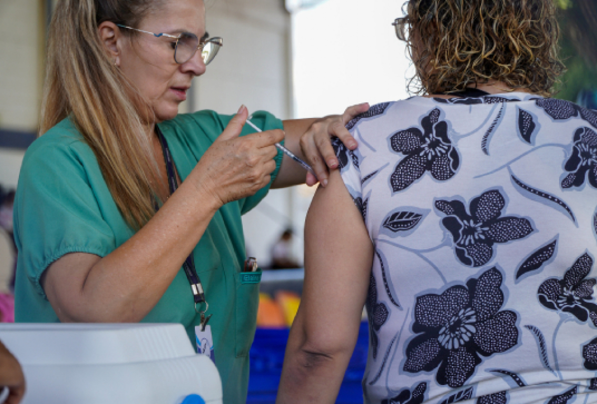 A imagem mostra uma pessoa em uniforme médico verde administrando uma vacina em outra pessoa.