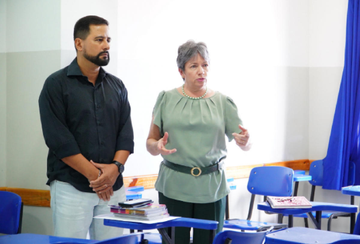 Duas pessoas conversam em uma sala de aula, com carteiras e materiais escolares ao redor.