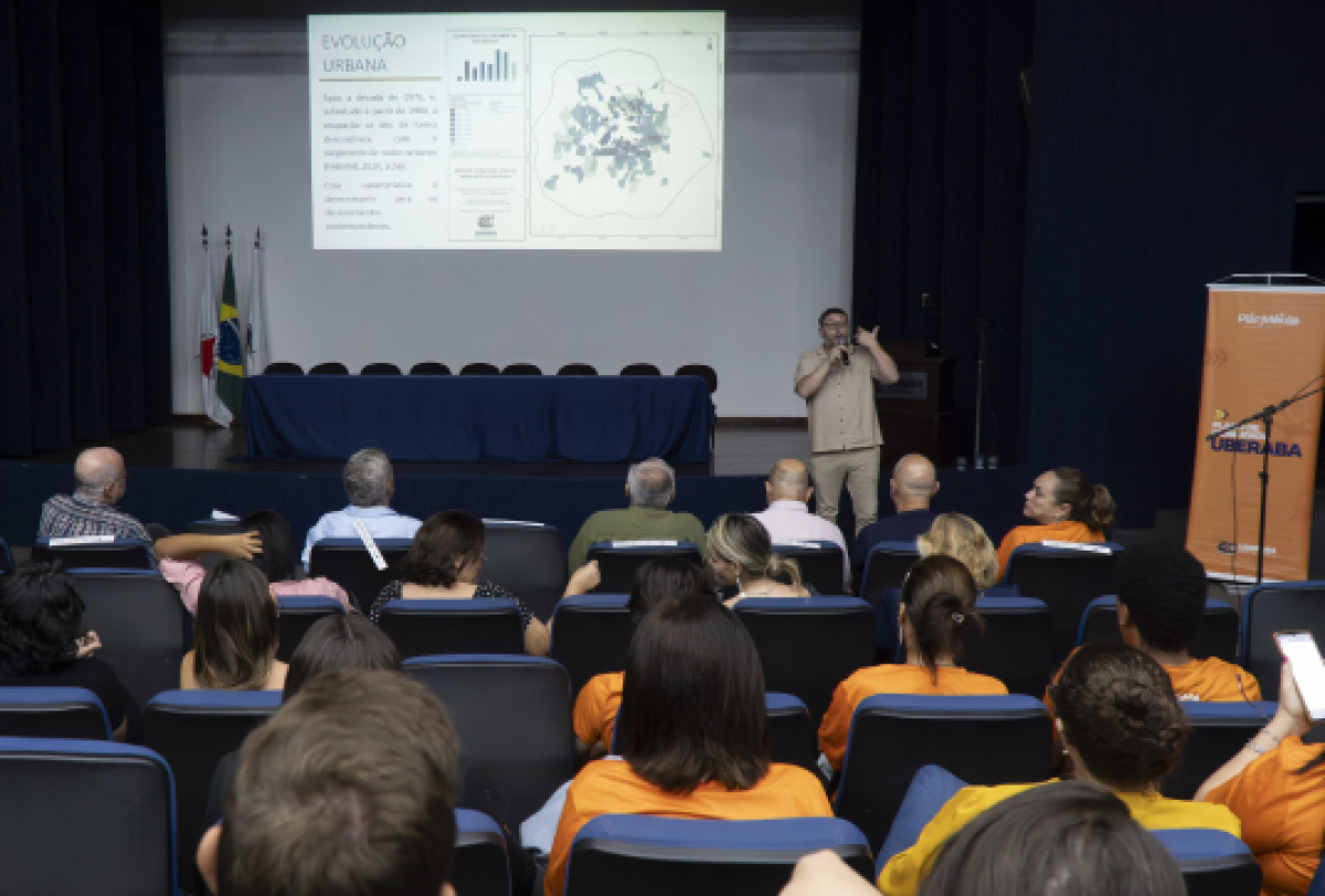 Auditório com pessoas assistindo uma  palestrante, ao fundo um telão.