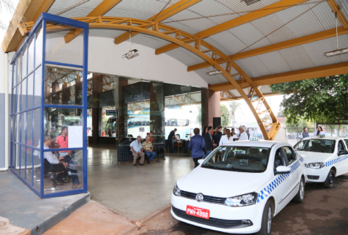 A imagem mostra um terminal rodoviário, com táxis estacionados na entrada e pessoas aguardando ou transitando pelo local. 