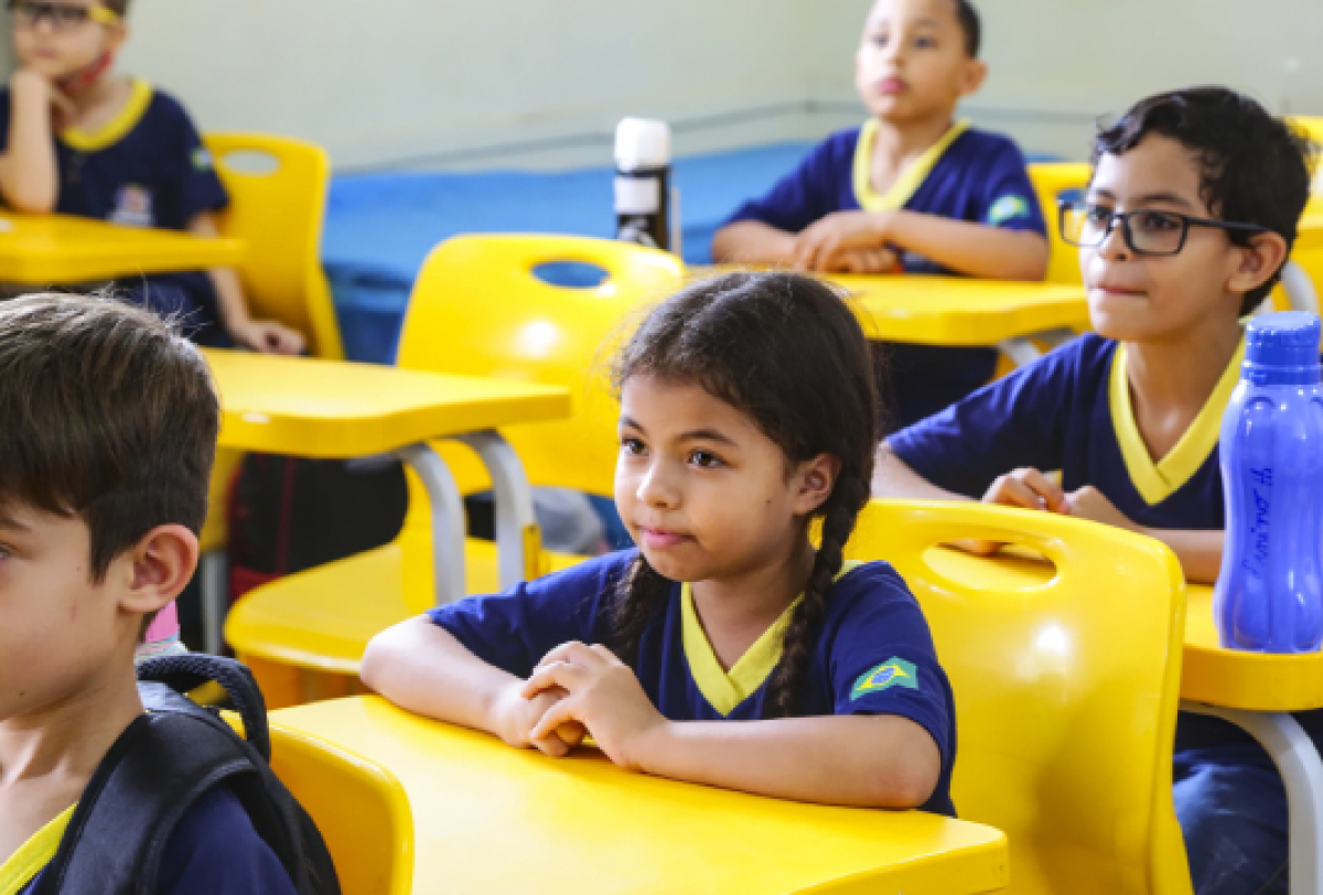 A imagem mostra crianças em uma sala de aula, sentadas em carteiras amarelas e vestindo uniformes escolares com detalhes na cor azul e amarela, incluindo uma bandeira do Brasil nas mangas. Elas parecem atentas e concentradas no que está sendo ensinado.