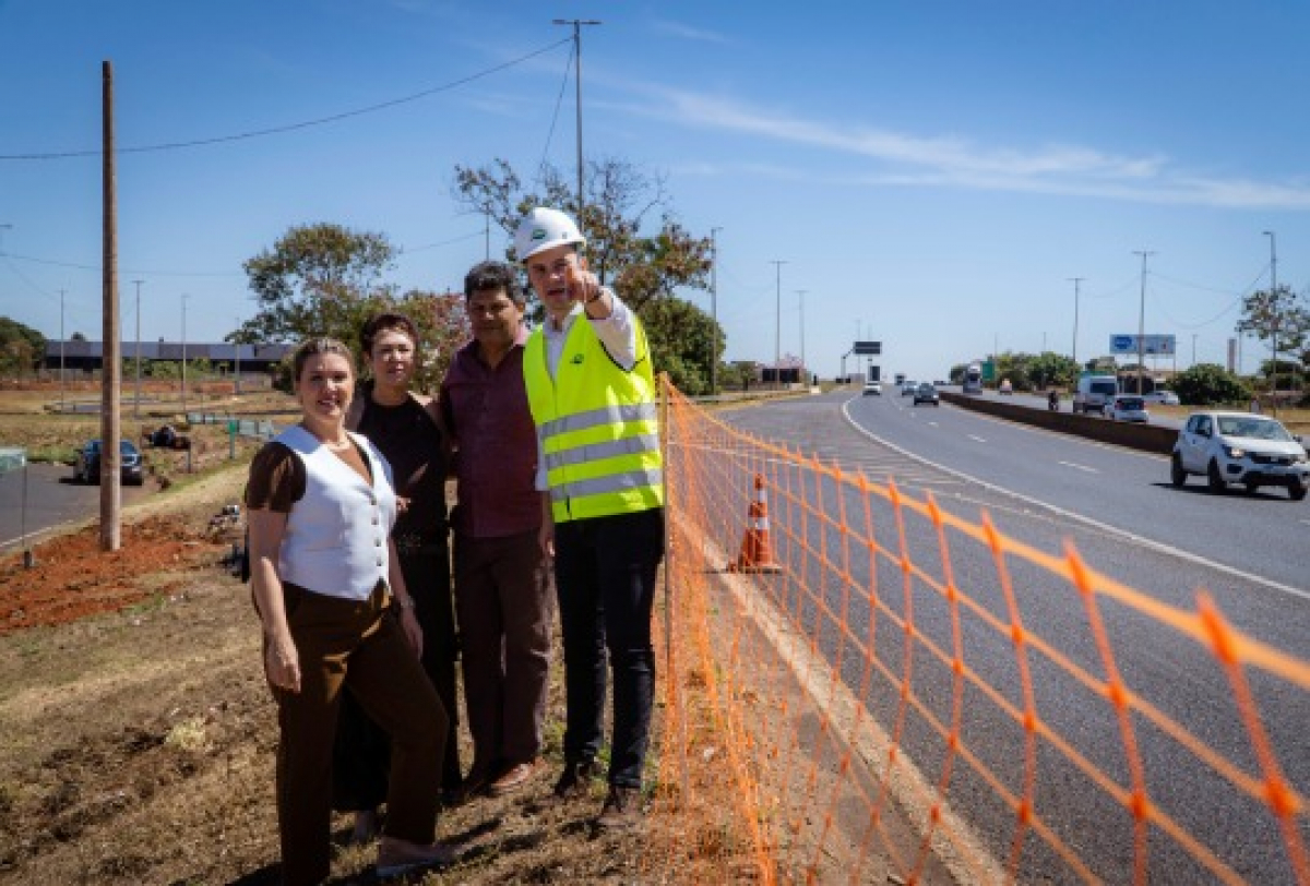 Grupo posa ao lado de rodovia em obra, com engenheiro apontando e tela de segurança laranja delimitando a área.