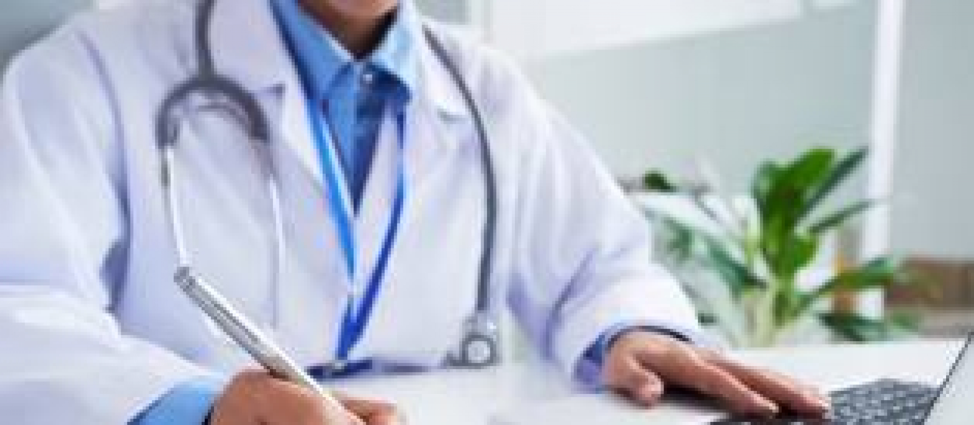 Médico em consultório escrevendo em uma prancheta e usando um laptop.