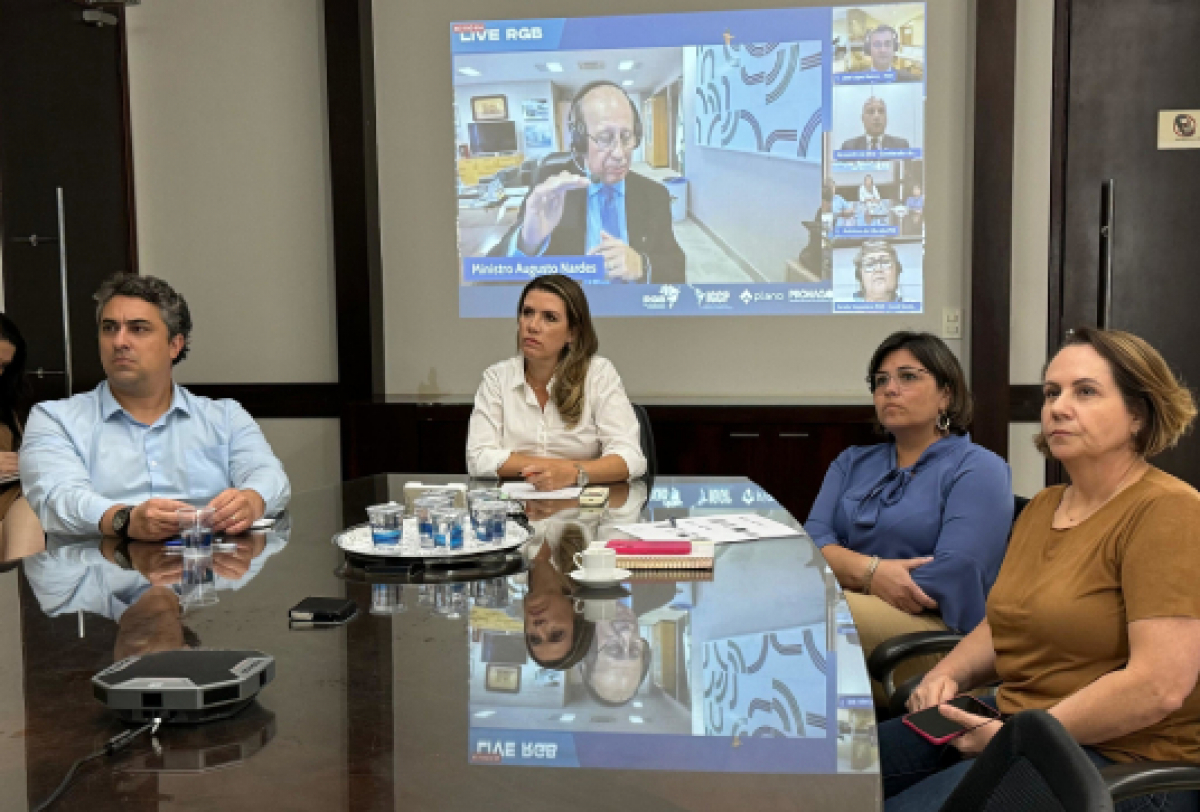 Reunião presencial com quatro pessoas em torno de uma mesa, acompanhando uma videoconferência projetada na parede.