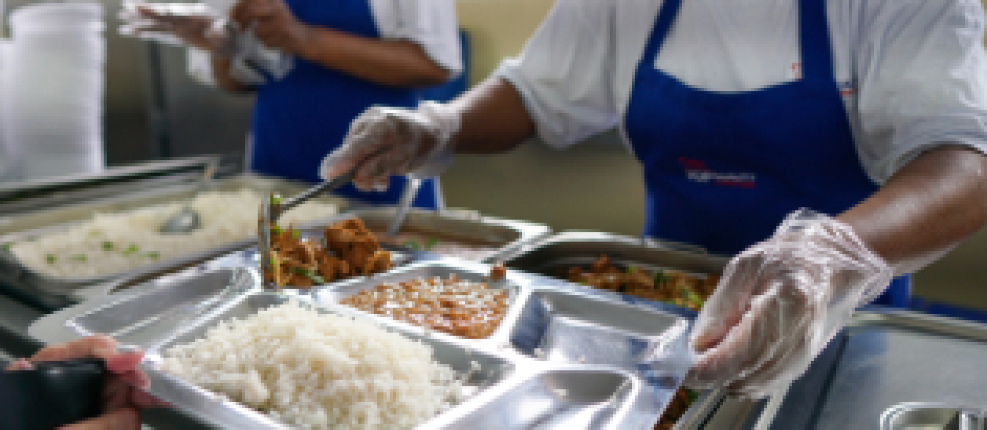 Distribuição de refeições em um restaurante popular, com atendentes servindo comida em bandejas de aço inox.