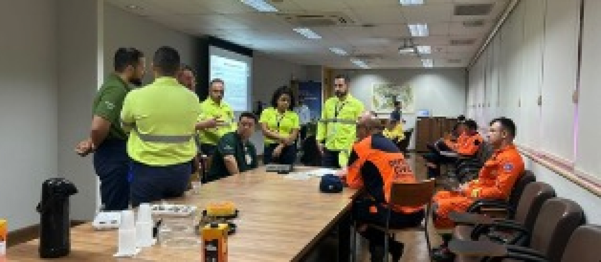 Reunião com equipes de emergência e defesa civil em uma sala, com apresentação projetada e materiais sobre a mesa.