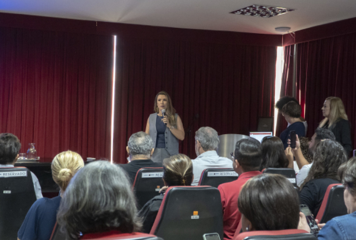 A imagem mostra um grupo de pessoas sentadas em uma sala, assistindo atentamente a uma pessoa que está de pé na frente, falando ou apresentando, em uma sala com cortinas vermelhas.