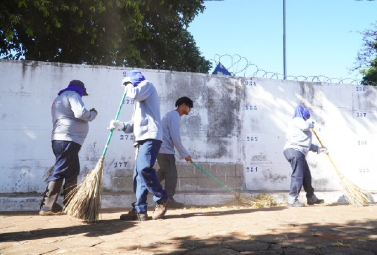 Equipe realiza limpeza urbana com varrição em calçada próxima a muro branco.