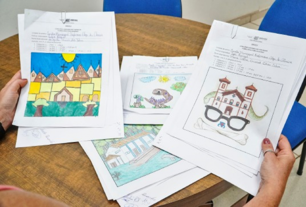 Pessoa segura desenhos coloridos de um concurso artístico, todos fixados em folhas com identificação oficial.