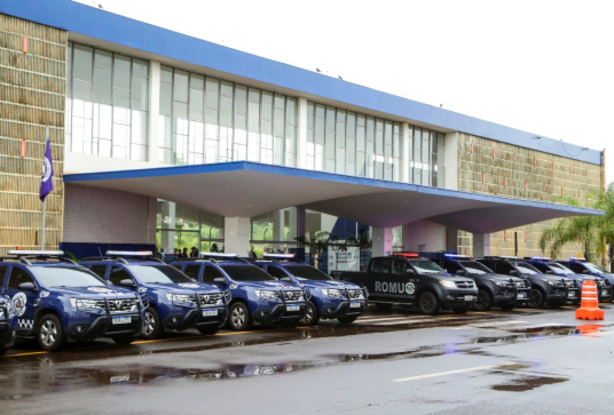 A imagem mostra uma fileira de veículos guarda municipal  estacionados em frente a um prédio moderno com telhado azul e grandes janelas de vidro.