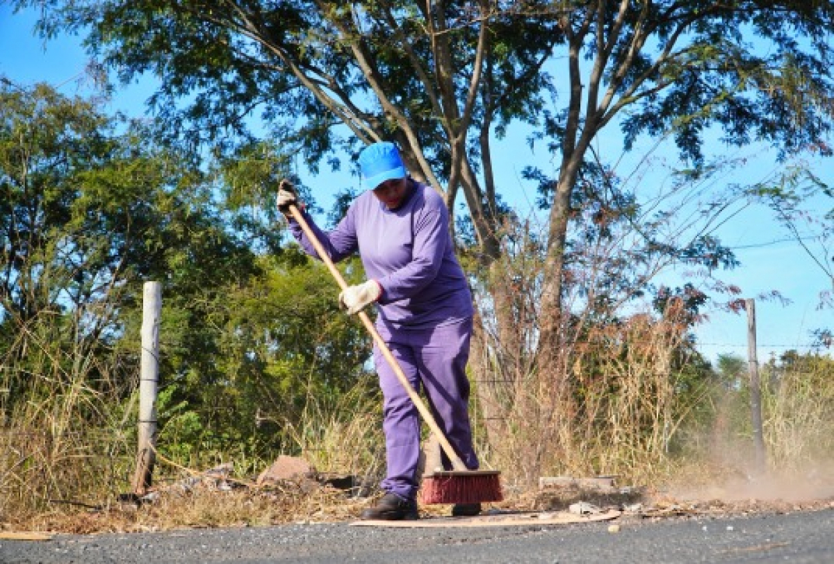 Servidor realiza varrição e limpeza da margem de uma via asfaltada.