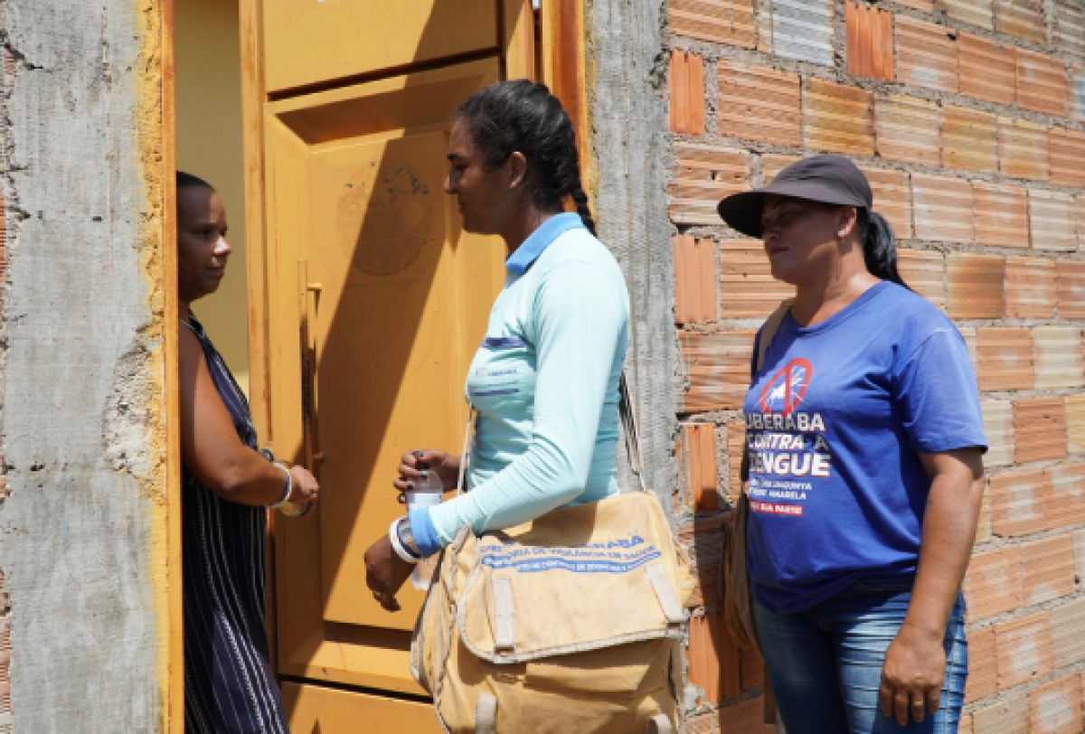 A imagem mostra três pessoas em pé na entrada de um prédio inacabado, sugerindo envolvimento em uma campanha de saúde pública, relacionada à prevenção da dengue. 