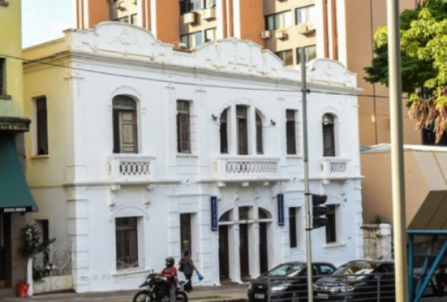 Imagem mostra um edifício branco de dois andares com fachada ornamentada, janelas arqueadas e elementos decorativos. Em frente, há veículos estacionados. Ao fundo, há edifícios modernos mais alto. 