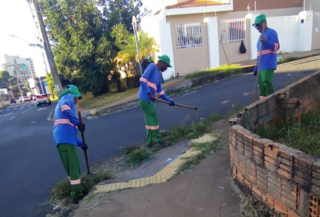 Três trabalhadores municipais realizam a limpeza e capina de calçada em área urbana.