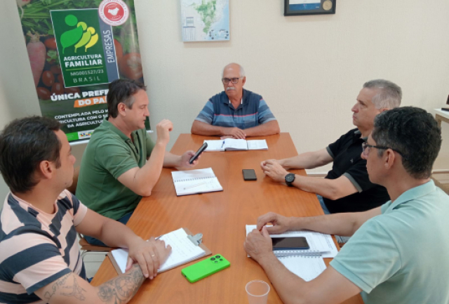 Uma reunião de cinco homens discutindo documentos em uma mesa, com um banner sobre agricultura