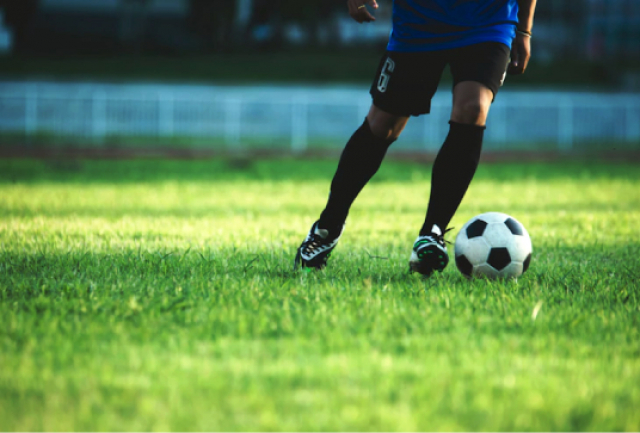 Jogador de futebol em campo, conduzindo a bola com o pé próximo ao gramado.