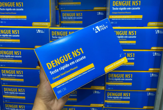 Uma mão segurando uma caixa de teste rápido de dengue NS1, mostrando um grande suprimento desses kits.