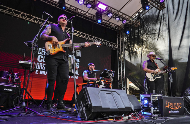 Banda se apresenta em palco ao ar livre durante evento musical, com guitarristas e baterista animando o público.