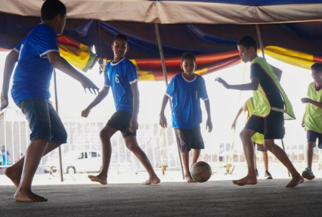 Meninos jogam futebol descalços em um espaço coberto, divididos em times de azul e verde.