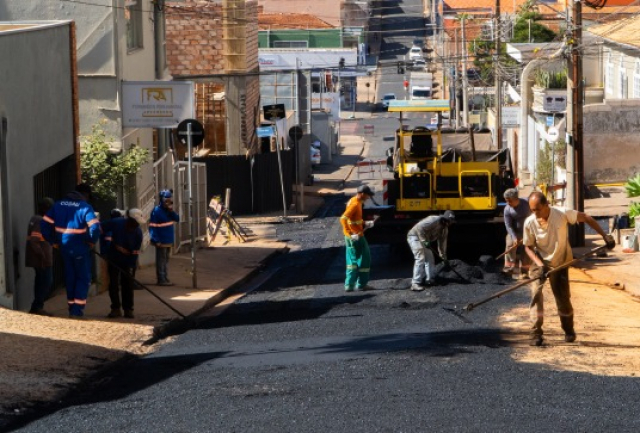 Equipe realiza obra de pavimentação asfáltica em rua urbana com auxílio de maquinário.