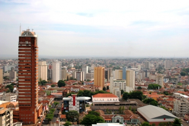 A imagem mostra uma paisagem urbana com uma mistura de edifícios altos e menores, com destaque para um prédio de tijolos vermelhos à esquerda e um céu claro com algumas nuvens.