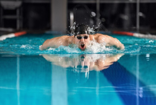 A imagem mostra um nadador executando a braçada borboleta em uma piscina, com água espirrando ao redor. A piscina tem uma marca azul de pista visível na água.